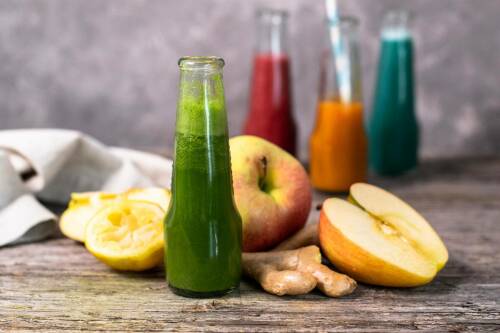 Grüner Ingwer Shot mit Spinat in Flaschen neben Äpfeln und Zitrone vor hellem Hintergrund.