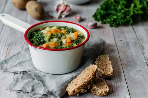 Grünkohleintopf in Pfännchen neben Vollkornbrot und Kartoffeln vor hellem Hintergrund.