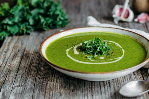 Grünkohlsuppe in Teller neben Grünkohl und Knoblauch vor hellem Hintergrund.