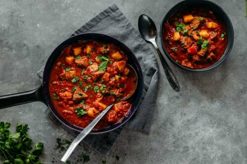 Gulaschsuppe auf schwarzen Tellern neben Besteck und Koriander vor dunklem Hintergrund.