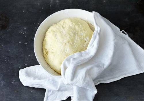 Weiße Schüssel gefüllt mir Hefeteig, noch halb mit einem Geschirrtuch abgedeckt, auf einer schwarzen Arbeitsplatte. Von oben fotografiert.
