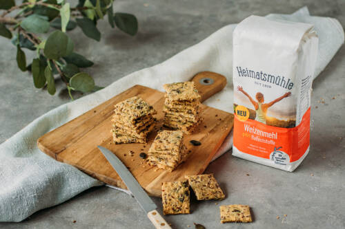 Selbstgemachtes Knäckebrot in kleinen Quadraten auf einem Holzbrett gestapelt, daneben eine Packung "Weizenmehl plus Ballaststoffe" von Heimatsmühle