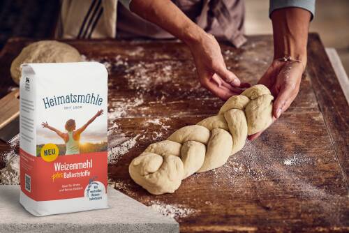 Ein Hefezopf wird auf einem Holztisch geflochten, daneben eine Packung "Weizenmehl plus Ballaststoffe" von Heimatsmühle