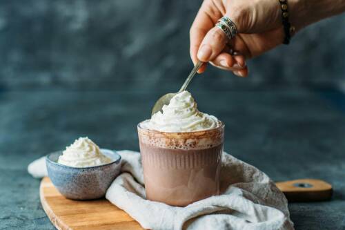 Heiße dunkle Schokolade mit Sahne in Glas und Hand mit Löffel vor dunklem Hintergrund.