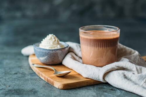Heiße dunkle Schokolade in Glas neben Schlagsahne und Löffel vor dunklem Hintergrund.