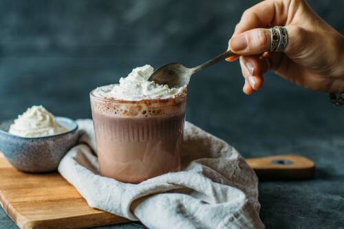 Heiße Schokolade in Glas mit Sahneklecks und Hand mit Löffel vor dunklem Hintergrund.