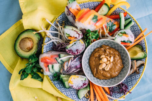 Teller mit buntem Gemüse, gefüllten Reispapierröllchen und Erdnussdip von oben fotografiert.