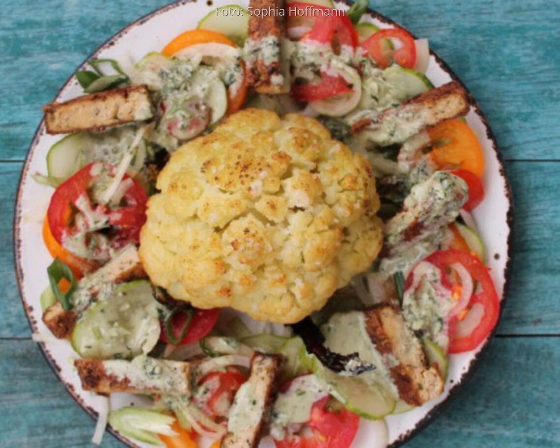 blaues Holzbrett und Teller mit ganzem gebackenen Blumenkohl und Gemüse