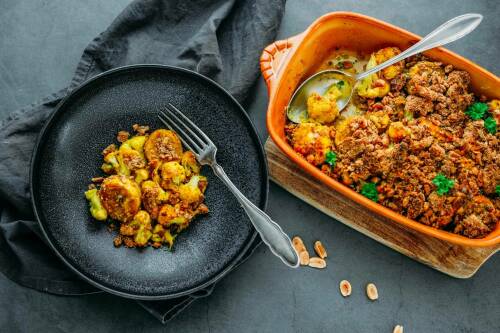 Indischer Blumenkohlauflauf in einer Auflaufform, daneben eine Portion auf einem schwarzen Teller.