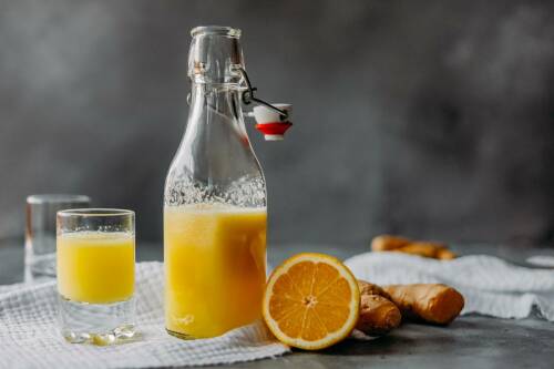 Ingwer Shot in Glas und Flasche neben Orange und Ingwer vor dunklem Hintergrund.