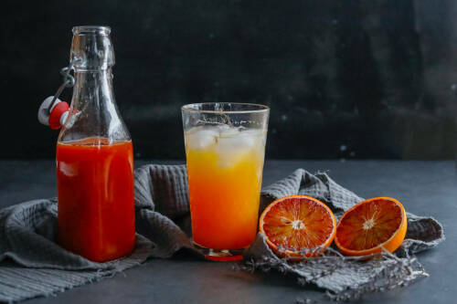 Ingwersirup mit Blutorange und Thymian in Glas und Flasche neben Blutorange vor dunklem Hintergrund.