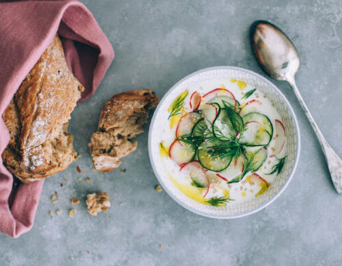 Joghurt griechischer Art in Schüssel neben Löffel und Brotlaib vor hellem Hintergrund.