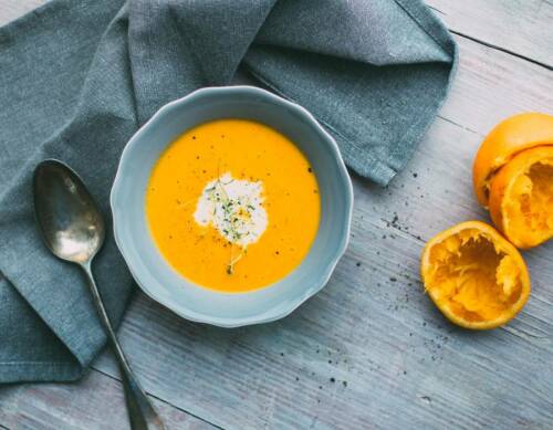 Karotten-Ingwer-Suppe in blauer Schüssel neben ausgedrückten Orangen vor dunklem Hintergrund.