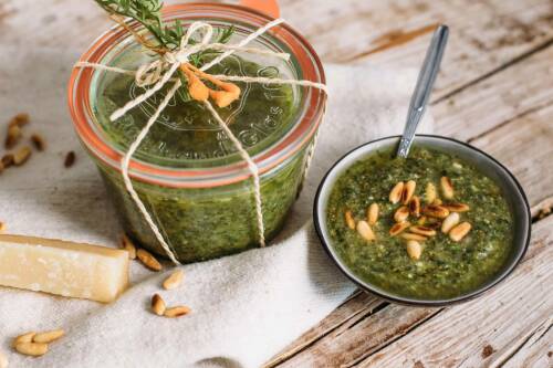 Karottengrün-Pesto in Schale und Weckglas mit Rosmarinzweig neben Parmesan und Pinienkernen auf Holstisch.