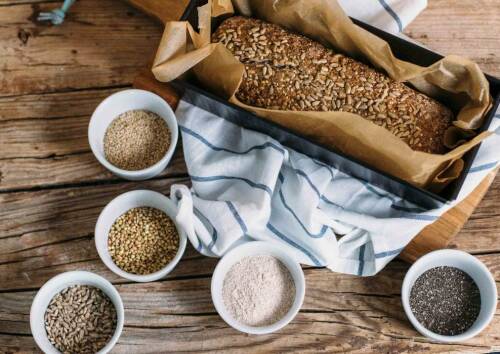 Körniges Kastenbrot, noch in der Form. Daneben verschiedene Samen und Körner. Von oben fotografiert.