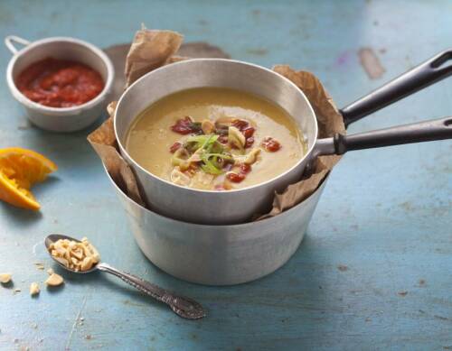 Kichererbsen-Erdnuss-Suppe in silbernem Topf neben Tomatensoße vor blauem Hintergrund.