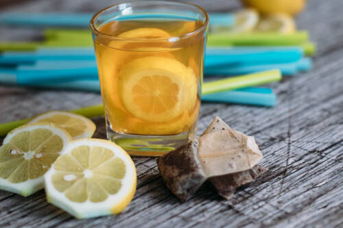 Glas Eistee aus Schwarztee, daneben Zitronenscheiben und Teebeutel, im Hintergrund Strohhalme, von vorne fotografiert