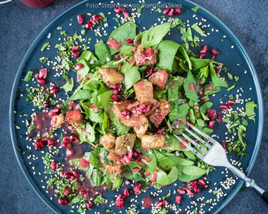 Dunkler Teller mit Babyspinat, Granatapfel und panierten Auberginenstücken.