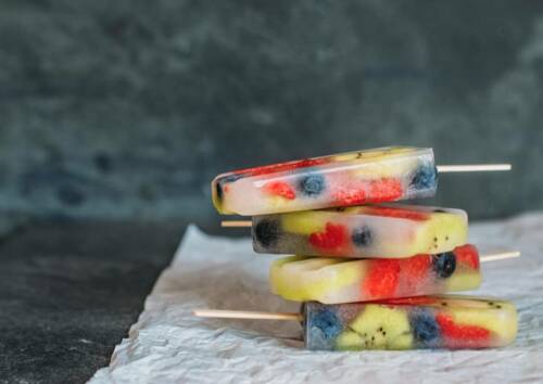 Vier Popsicles am Stiel gestapelt in der rechten unteren Bildecke mit Blaubeeren, Erdbeeren und Kiwi vor dunkelgrauem Hintergrund. Von vorne fotografiert.