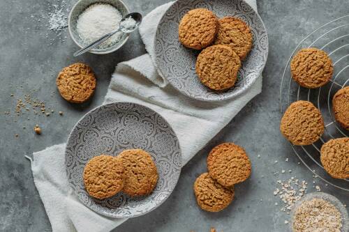 Kokos-Haferflocken-Kekse auf Tellern und Gitter neben Kokos- und Haferflocken vor hellem Hintergrund.