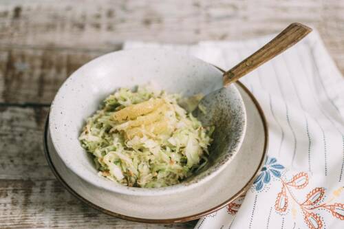 Krautsalat in weißer Schüssel mit Gabel auf Holztisch und Geschirrtuch vor hellem Hintergrund.