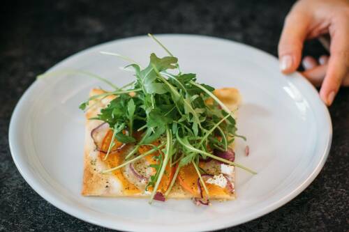 Kürbis Flammkuchen mit Rucola Haube auf weißem Teller mit Hand vor dunklem Hintergrund.