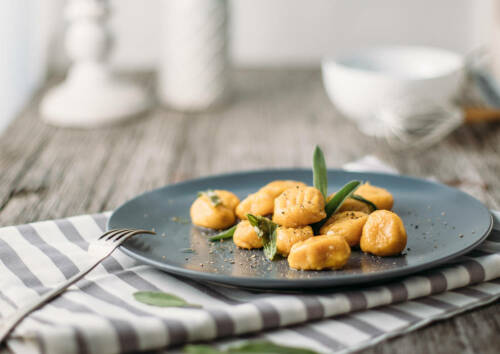 Orangene Kürbis-Gnocchi auf einem grauen Teller mit Salbeiblättern darauf, von der Seite fotografiert.