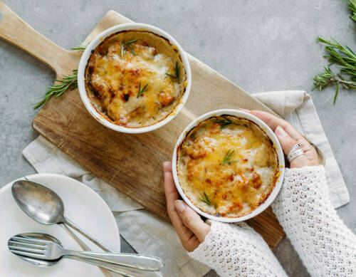 Kürbis-Graupen-Auflauf in zwei Schüsseln auf Schneidebrett mit Händen vor hellem Hintergrund.
