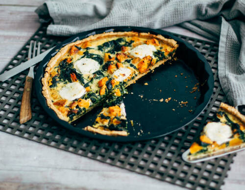 Kürbis-Spinat-Quiche auf Pizzablech mit Besteck vor dunklem Hintergrund.