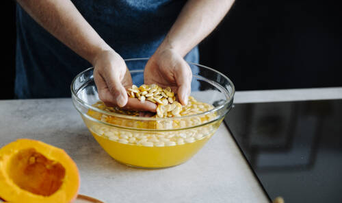 Kürbiskerne in Schale mit Wasser und Händen vor dunklem Hintergrund.