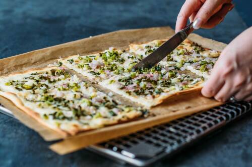 Lauchzwiebel Flammkuchen auf Backblech mit Hand und Messer vor dunklem Hintergrund.
