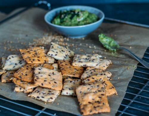 Leinsamen-Dinkel-Cracker auf Backblech neben Pesto vor dunklem Hintergrund.