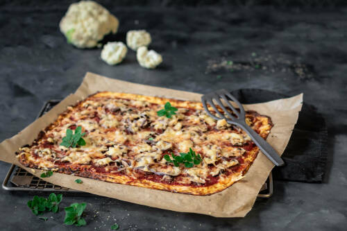 Low Carb Blumenkohl Pizza auf Backbleck neben Blumenkohl vor dunklem Hintergrund.