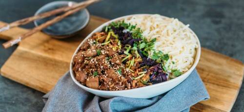 Teriyaki Bowl mit Jackfruit, Rotkohl, Karotten und Lauch in einer Schüssel. Dazu Mie-Nudeln, im Hintergrund Stäbchen auf einer Schüssel.