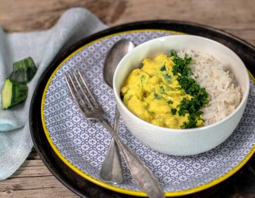 Mandel-Zucchini-Curry in Schüssel mit Besteck auf Teller vor hellem Hintergrund.