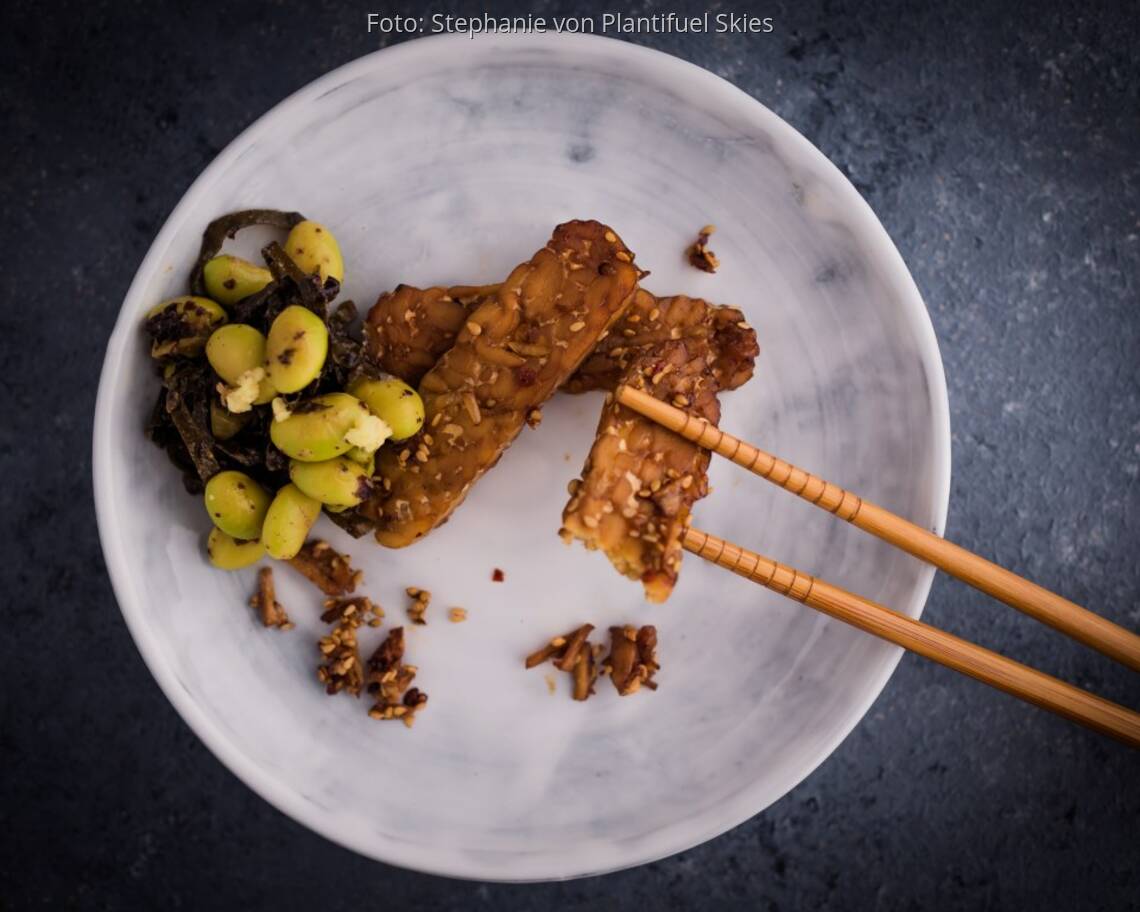 Weißer Teller mit mariniertem Tempeh und Edamame, dazu Essstäbchen auf dunkler Arbeitsplatte