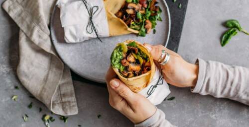 Pfannkuchen Wraps gefüllt mit Spinat, Pilzen und Staudensellerie auf einem grauen Teller vor grauem Hintergrund. Zu sehen ist ein weiterer Pfannkuchen Wrap der von zwei Händen gehalten wird, von oben fotografiert.