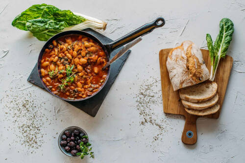 Mediterraner Bohnen-Mangold-Eintopf in Pfanne neben Hausbrot vor hellem Hintergrund.