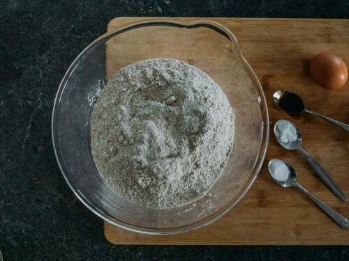 Mehl und Zucker neben Ei auf Schneidebrett vor dunklem Hintergrund.