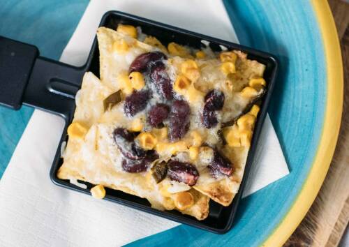 Mexikanisches Raclette-Pfännchen mit Bohnen, Tacos und Mais mit Käse überbacken. Von oben fotografiert.