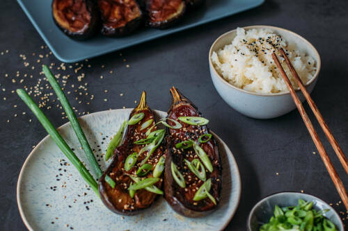 Miso Auberginen mit Sesam und Frühlingszwiebeln auf Teller neben Basmatireis und Stäbchen.
