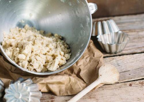 Mürbteig in einer Rührschüssel, daneben Kochlöffel und Törtchen-Formen. Von vorne fotografiert.