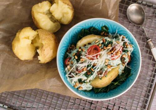Ofenkartoffel gefüllt mit Spinat-Ricotta-Sauce, dazu Pinienkerne und Tomaten. Von oben fotografiert auf einem Gitter, daneben ein Löffel.
