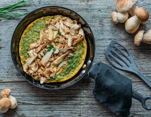 Omelett mit Steinpilzen in Pfanne neben Steinpilzen vor dunklem Hintergrund.