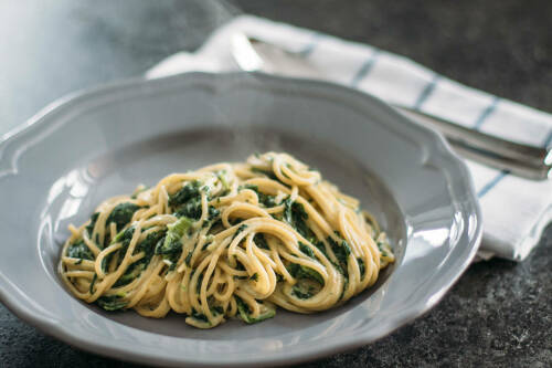One Pot Pasta mit Spinat in grauem Teller neben Geschirrtuch vor dunklem Hintergrund.