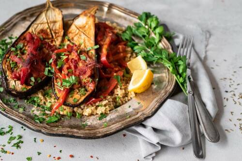 Orientalische Ofen-Auberginen auf Silberteller mit Besteck und Koriander vor hellem Hintergrund.