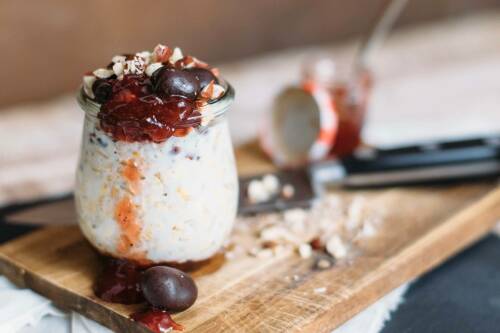 Overnight Oats mit Zwetschgen und Mandeln in Glas auf Schneidebrett mit Messer vor hellem Hintergrund.