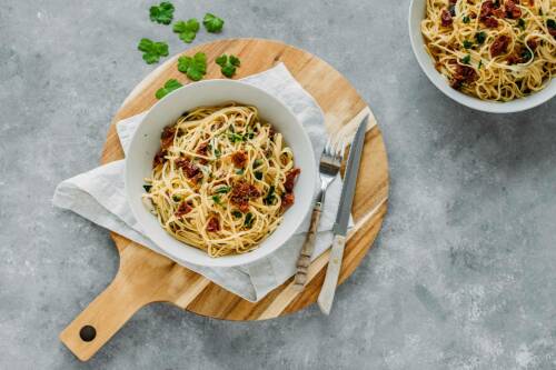 Pasta mit getrockneten Tomaten und Mozzarella auf weißem Teller und Brotzeitbrett vor hellem Hintergrund.