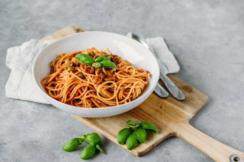 Pasta mit Oliven und Kapern auf weißem Teller und Schneidebrett vor hellem Hintergrund.