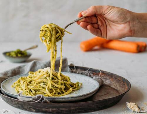 Pasta mit Pesto aus Karottengrün um Gabel gewickelt auf grauem Teller mit Hand vor hellem Hintergrund.